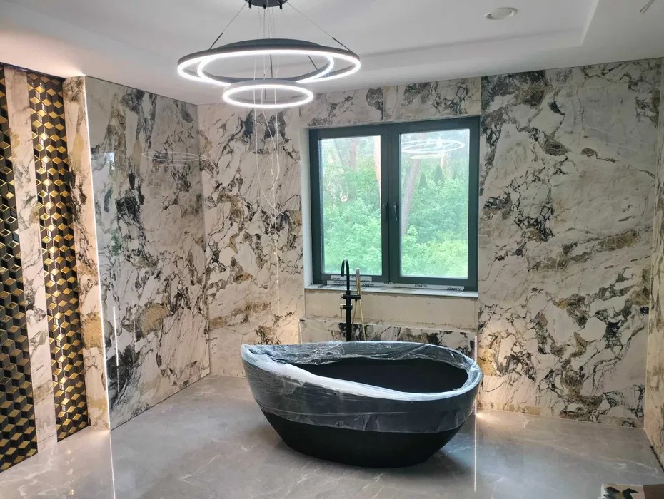 Elegancka łazienka w trakcie wykończenia: marmurowe ściany, czarna wanna zabezpieczona folią, nowoczesna lampa i okno z widokiem na zieleń.