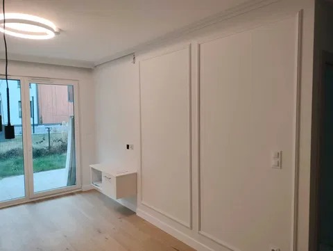 Jasne wnętrze po remoncie: białe ściany z dekoracyjnymi panelami, drewniana podłoga, przesuwne drzwi balkonowe i minimalistyczna szafka wisząca. Oświetlenie sufitowe.