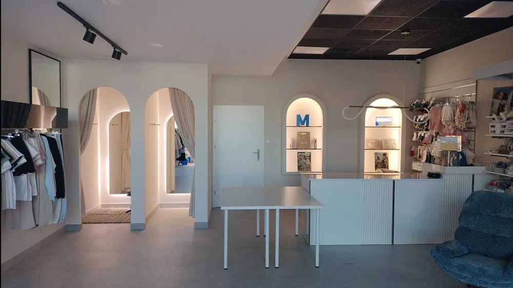 Jasne wnętrze lokalu usługowego z przymierzalniami w łukowych wnękach, podwieszanym sufitem i oświetleniem punktowym. Minimalistyczny design z białymi meblami i szarymi podłogami.