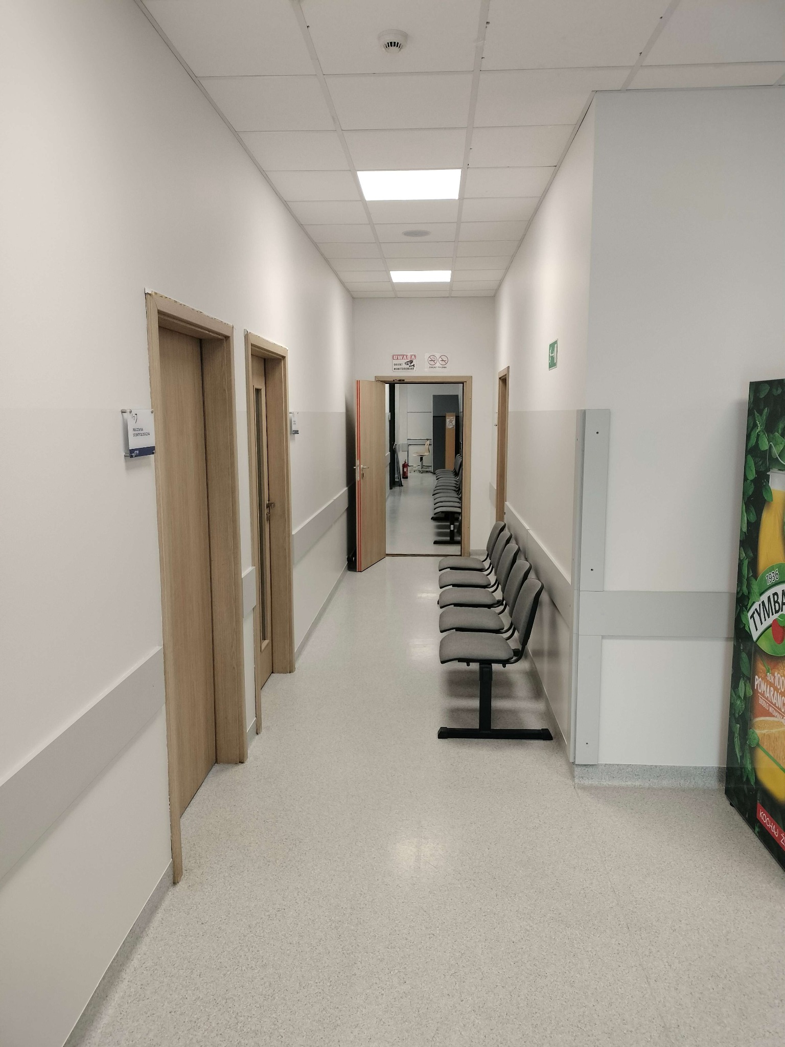 Korytarz w budynku szkoły po malowaniu ścian i uzupełnieniu gładzi. Widoczne drzwi, ściany pomalowane na biało, krzesła dla oczekujących i elementy identyfikacji wizualnej.
