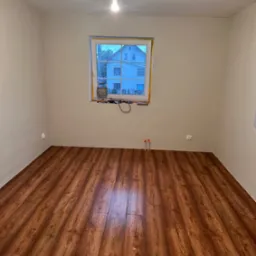 Pusty pokój po remoncie: świeżo położona podłoga z paneli imitujących drewno, pomalowane ściany na jasny kolor, okno z narzędziami na parapecie.