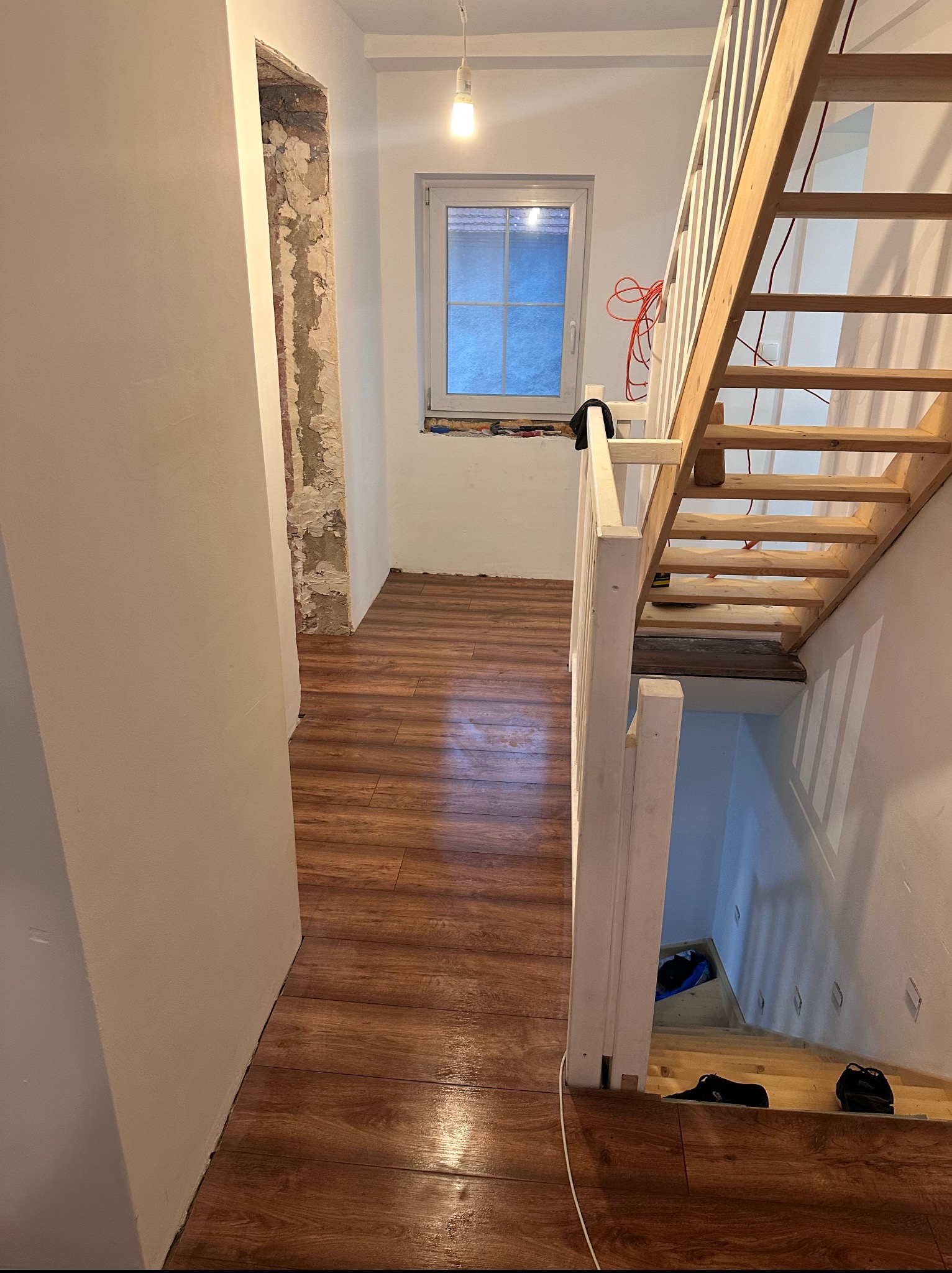 Wnętrze w trakcie remontu: surowe ściany obok gładkich, drewniana podłoga, schody z surowego drewna i instalacja elektryczna. Widok na okno i fragment korytarza.
