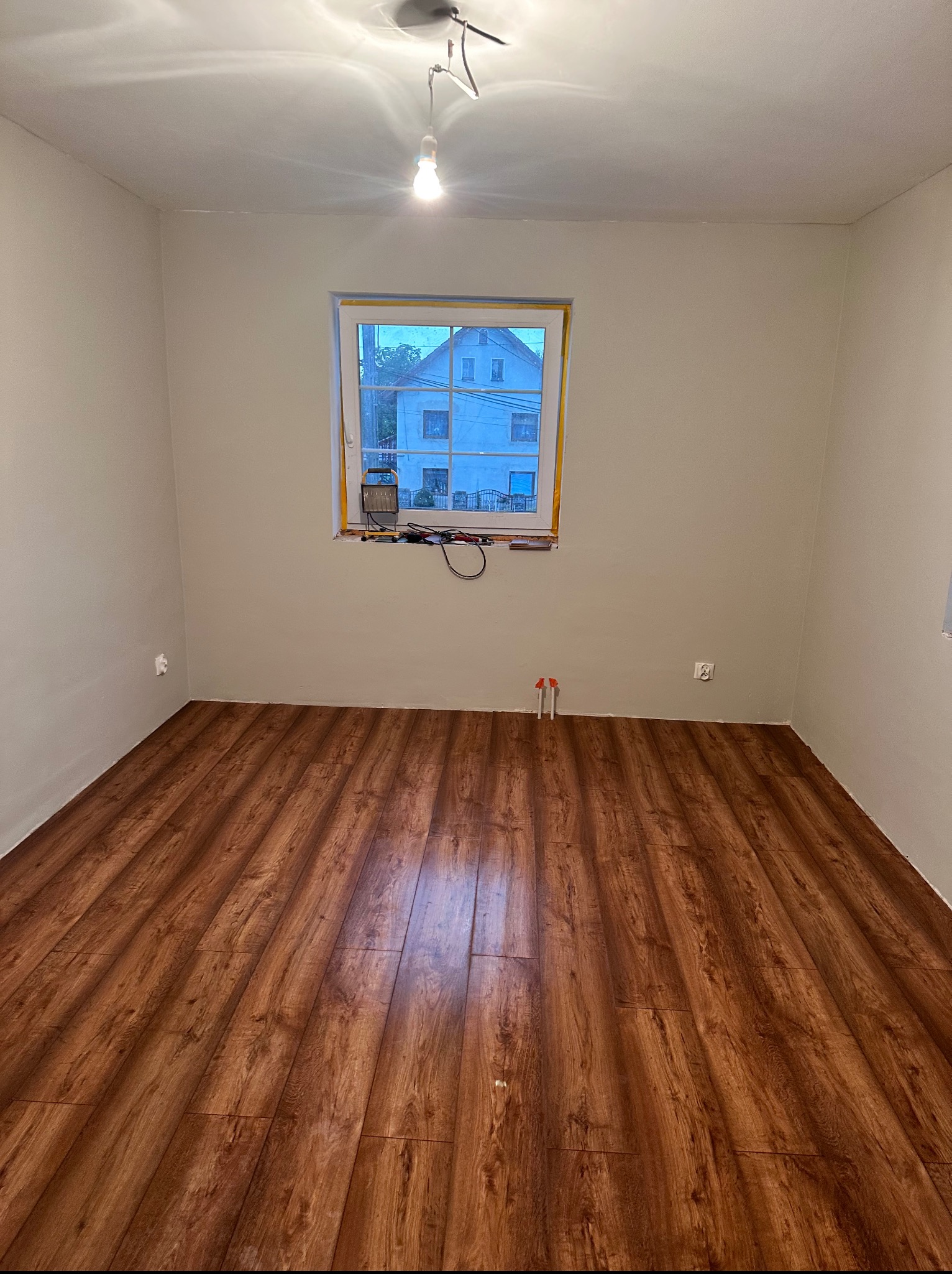 Pusty pokój po remoncie: świeżo położona podłoga z paneli imitujących drewno, pomalowane ściany na jasny kolor, okno z narzędziami na parapecie.