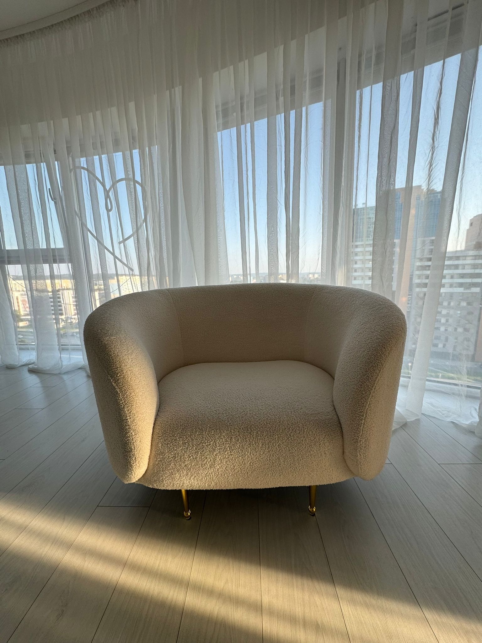 Kremowy fotel typu teddy na złotych nóżkach, na tle okna z firanką i widokiem na miasto. Na podłodze widoczne promienie słoneczne.