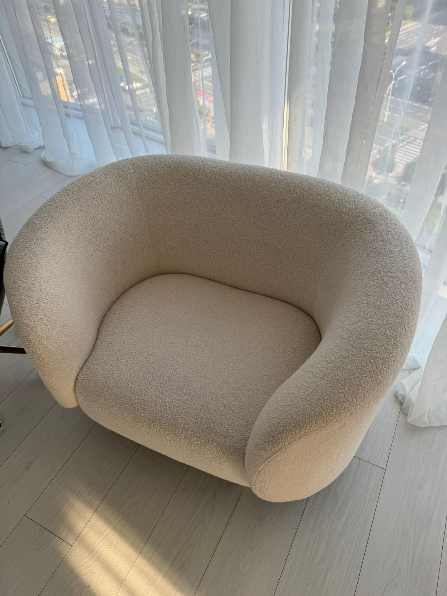 Kremowy fotel typu teddy bear na drewnianej podłodze, z delikatną zasłoną w tle. Widoczne subtelne światło słoneczne i miękka faktura tapicerki.