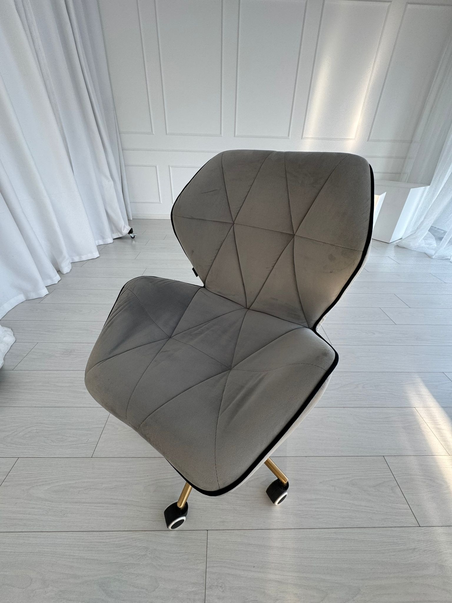 Szare, welurowe krzesło biurowe na kółkach z czarną lamówką, na tle białych ścian i podłogi, oświetlone naturalnym światłem. Widoczne delikatne plamy na siedzisku.