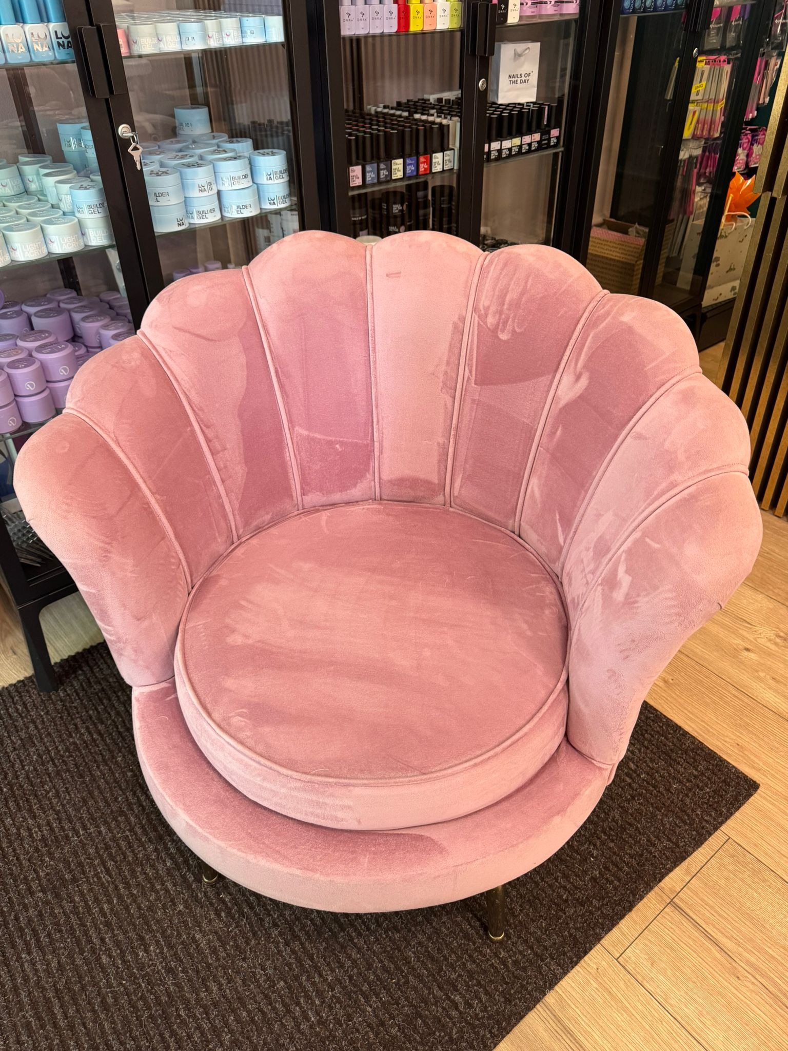 Różowy, pluszowy fotel w kształcie muszli na tle półek z kosmetykami do paznokci. Fotel czeka na pranie tapicerki, widoczne ślady użytkowania.