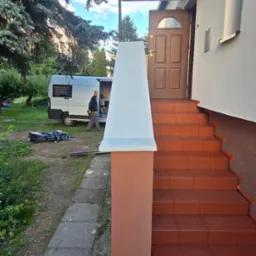 Odnowione schody wejściowe do domu z brązowymi płytkami i pomalowanymi filarami. W tle zaparkowany bus i osoba przy nim.