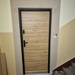 EMSI ZŁOTA RĄCZKA - Zamontowane drzwi wejściowe z czarną ramą i drewnopodobnym wypełnieniem w klatce schodowej w Inowrocławiu. Widoczne wykończenia wokół ościeżnicy.
