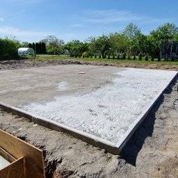 EMSI ZŁOTA RĄCZKA - Przygotowany teren pod budowę z wylaną podbudową, obrzeżami i wykopem. Widok na błękitne niebo i zieleń w tle. Ujęcie z dołu, z widoczną ręką trzymającą paczkę papierosów.