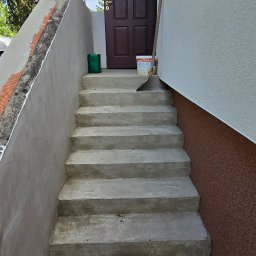EMSI ZŁOTA RĄCZKA - Betonowe schody wejściowe do domu po remoncie, z widoczną siatką wzmacniającą na ścianie i narzędziami malarskimi przy drzwiach. Świeżo pomalowana elewacja.
