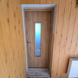 EMSI ZŁOTA RĄCZKA - Montaż drzwi wewnętrznych z szybą w obudowie z paneli ściennych. Widoczne wykończenia i fragment grzejnika. Ułożone płytki podłogowe.