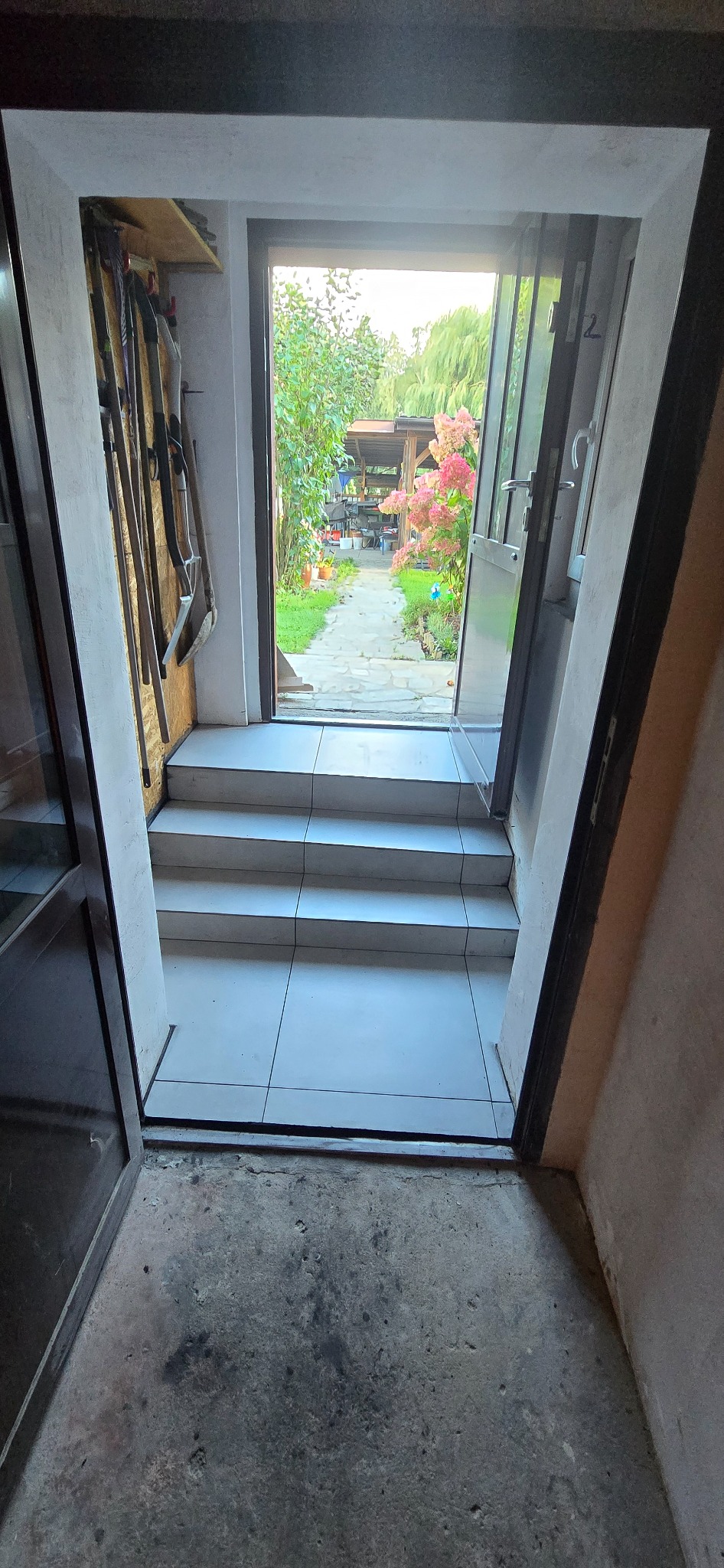 Wyjście na ogród przez przeszklone drzwi. Widoczne płytki na schodach i narzędzia ogrodnicze. W tle roślinność i altana.