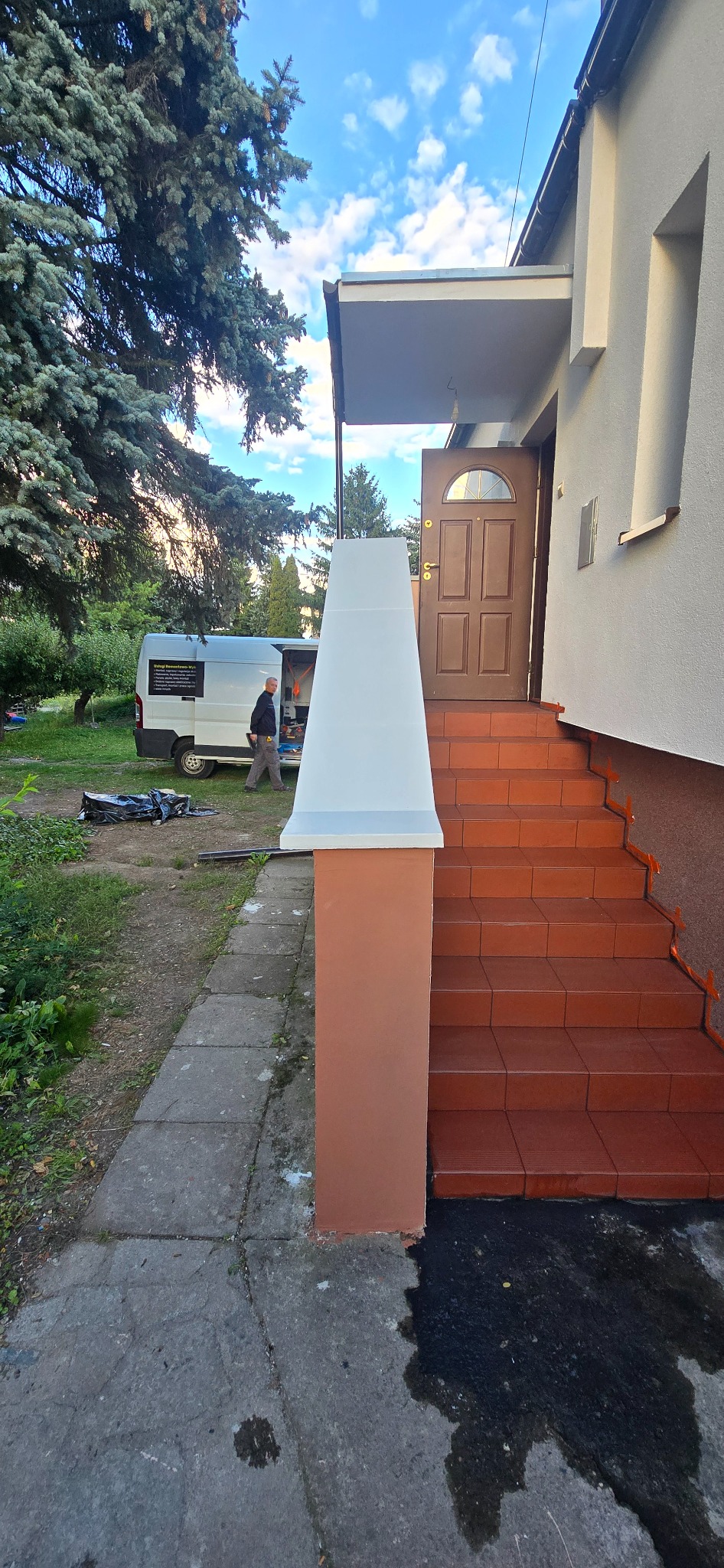 Odnowione schody wejściowe do domu z brązowymi płytkami i pomalowanymi filarami. W tle zaparkowany bus i osoba przy nim.
