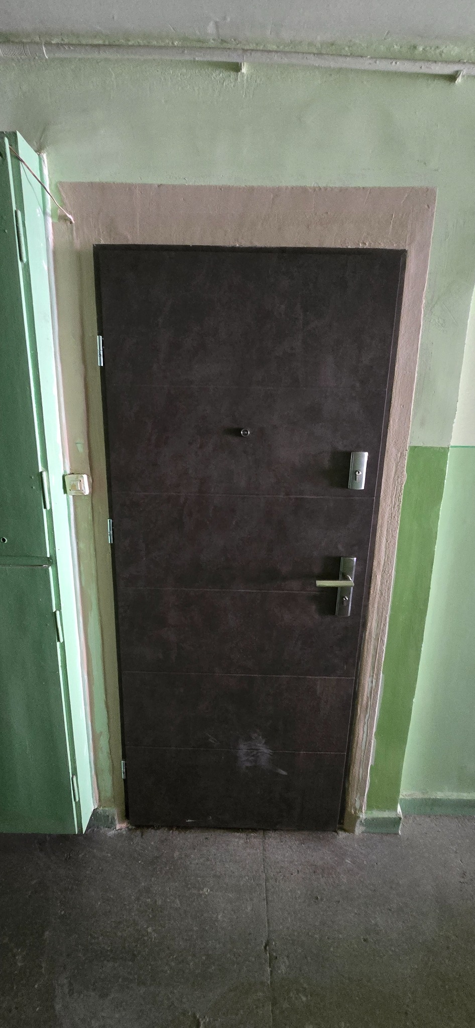 Ciemnobrązowe drzwi wejściowe z wizjerem i srebrną klamką, zamontowane w zielonej ścianie klatki schodowej. Widoczna ościeżnica i fragment sąsiednich, zielonych drzwi.