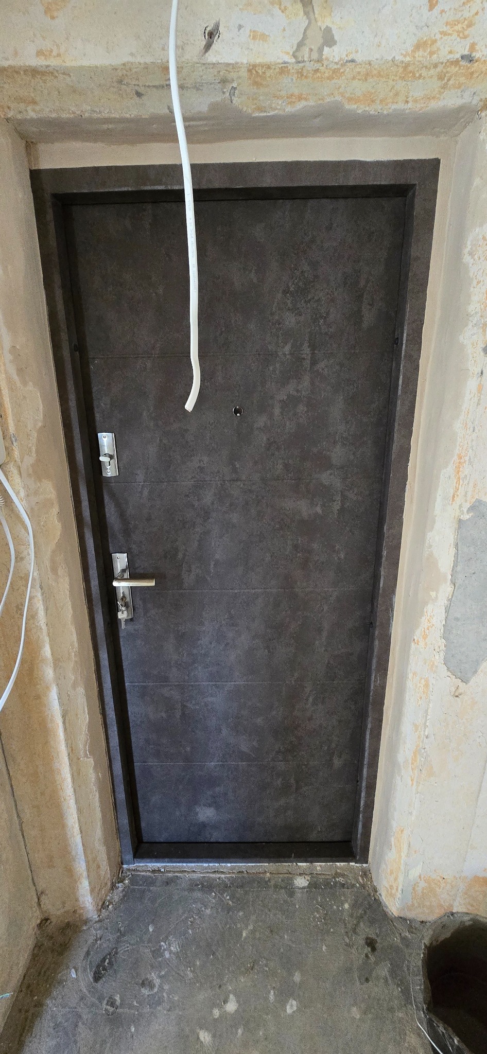 Ciemne, nowoczesne drzwi wejściowe z metalową klamką, zamontowane w surowym otworze ściany. Widoczne przewody elektryczne i niedokończone wykończenie.