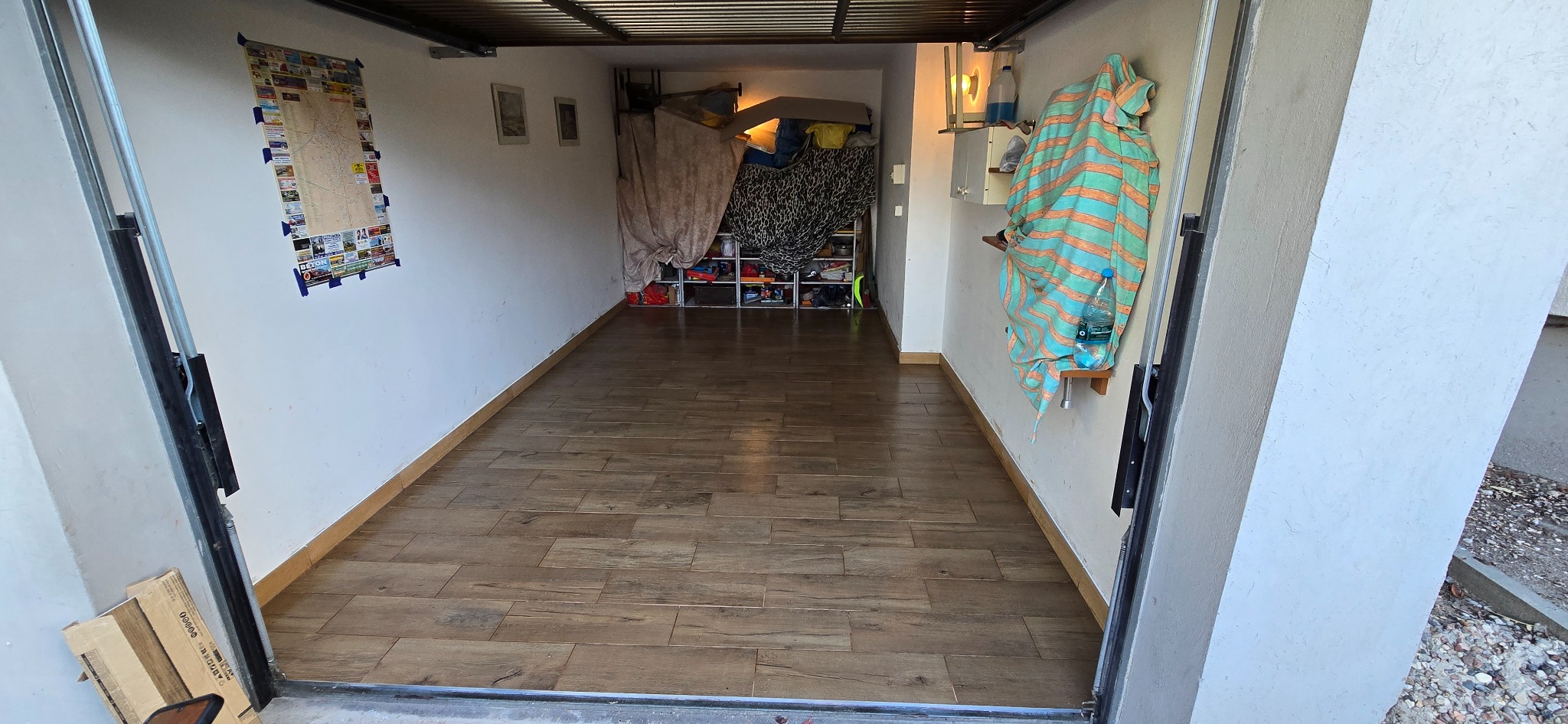 Wykończone wnętrze garażu z brązowymi panelami podłogowymi. Na ścianie wisi mapa i ręcznik. Widok z zewnątrz przez otwarte drzwi garażowe.