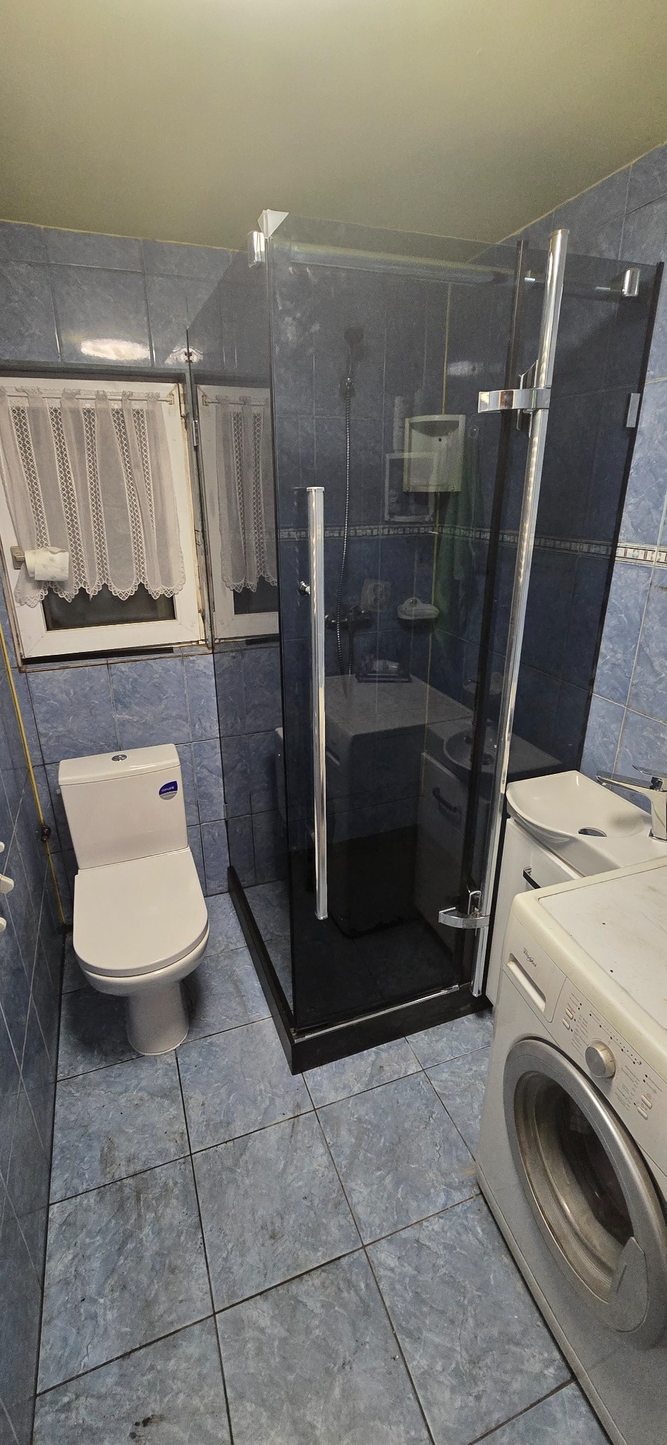 Niebieska łazienka po remoncie: kabina prysznicowa z ciemnego szkła, toaleta, umywalka i pralka. Widok na okno z firanką. Płytki podłogowe w odcieniach szarości.