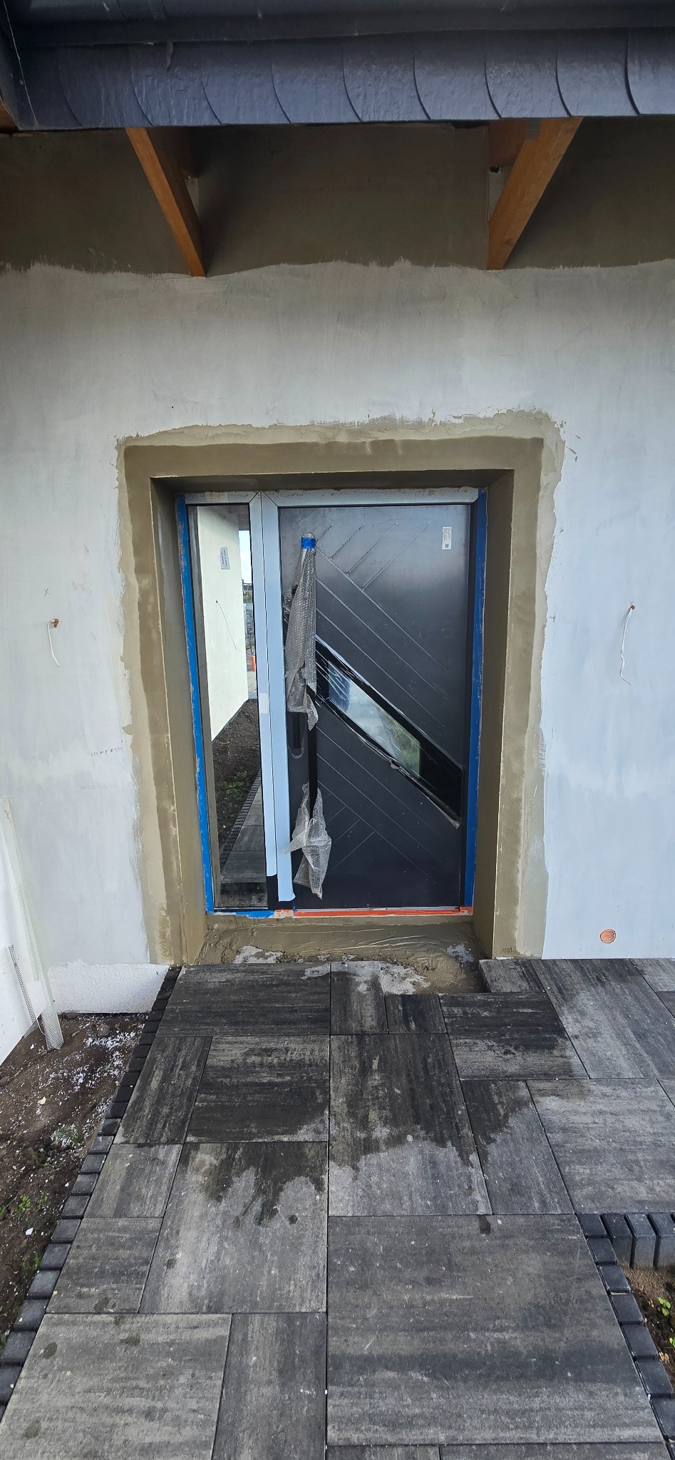 Montaż nowoczesnych, grafitowych drzwi wejściowych z przeszkleniem, osadzonych w surowym otworze z widocznymi śladami zaprawy i nową kostką brukową przed wejściem.