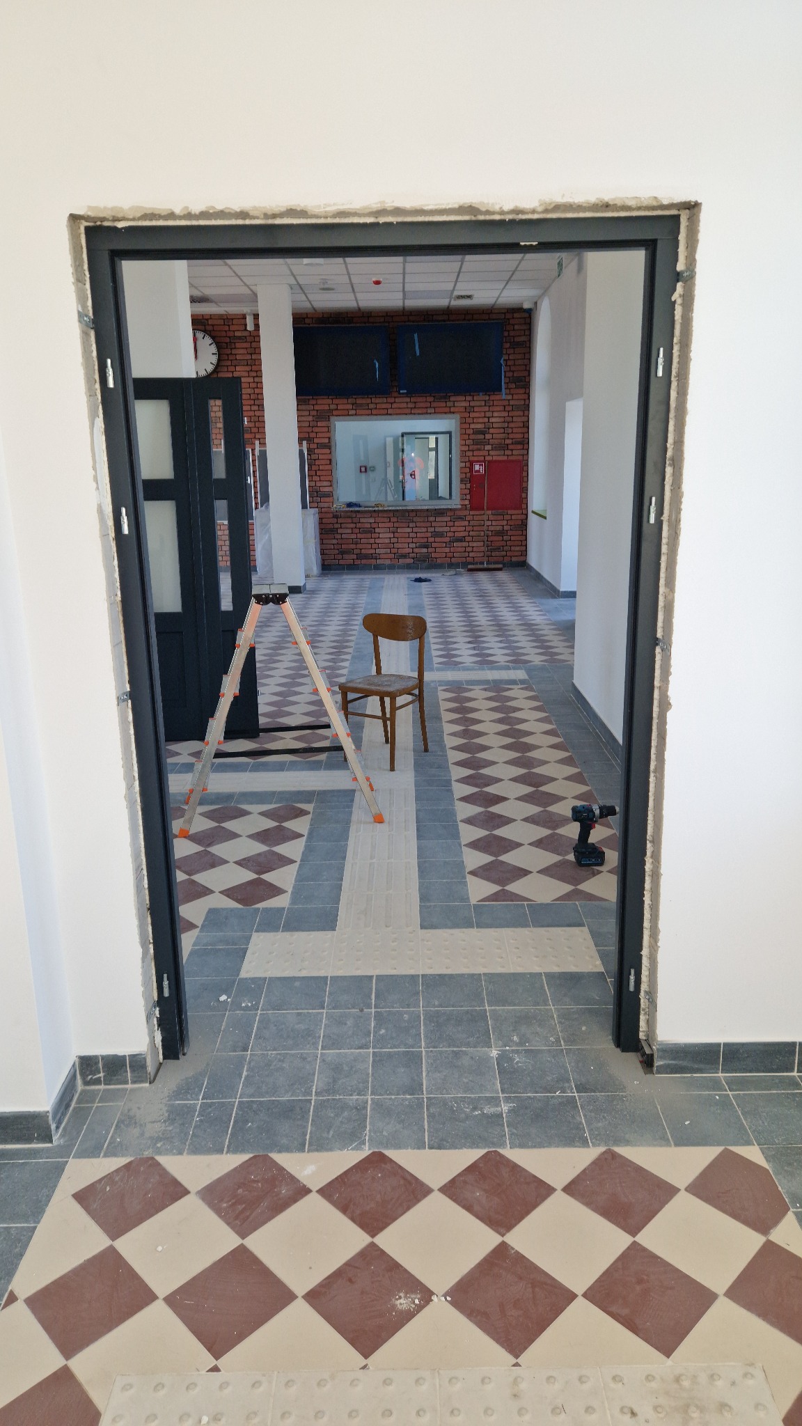 Montaż ościeżnicy w surowym otworze drzwiowym. W tle widoczna drabina, krzesło i narzędzia na podłodze z wzorzystą, częściowo ułożoną terakotą.