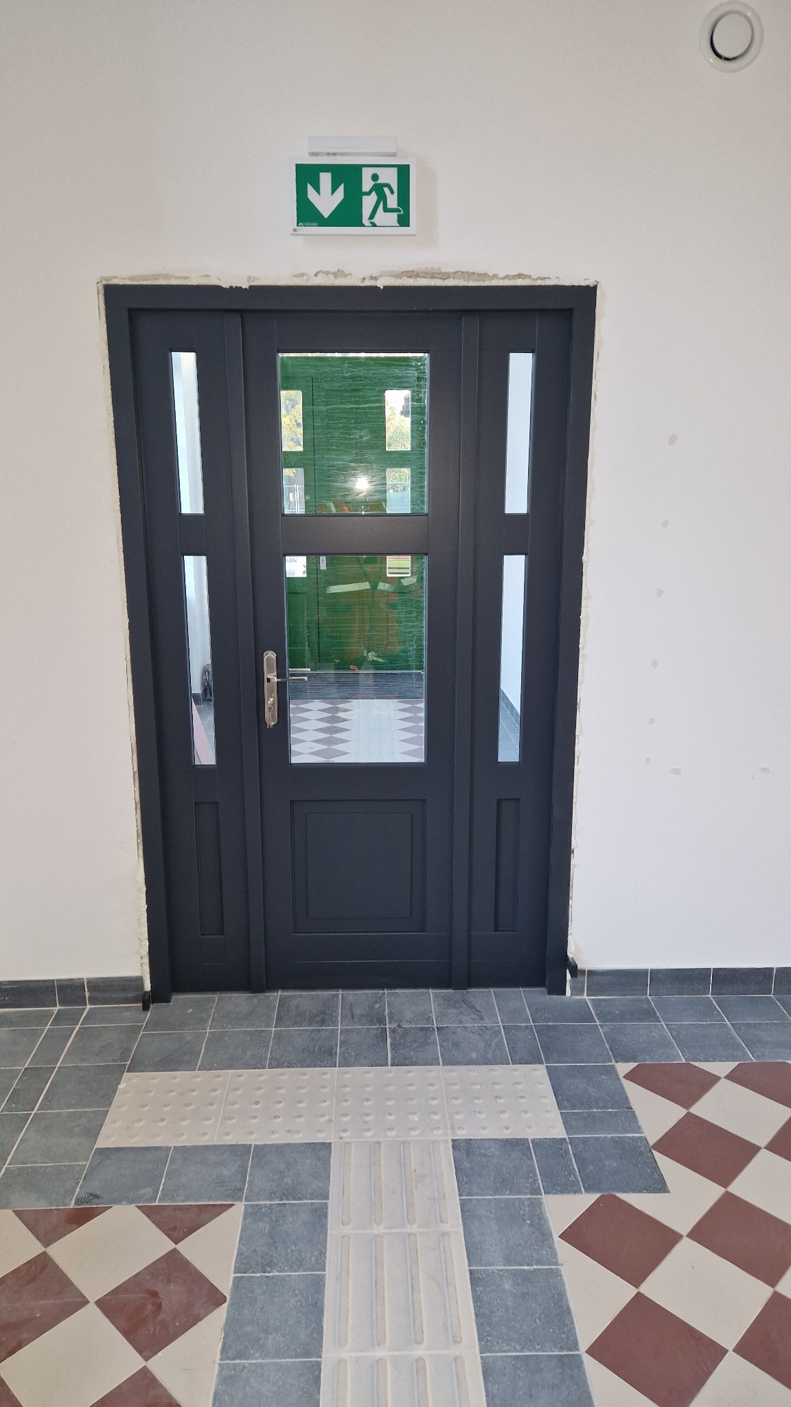 Ciemnoszare, przeszklone drzwi z oznaczeniem drogi ewakuacyjnej nad nimi, osadzone w białej ścianie, z widocznymi śladami po remoncie i płytkami podłogowymi.