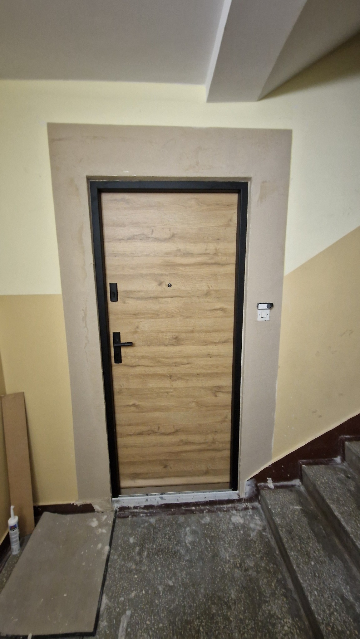 Zamontowane drzwi wejściowe z czarną ramą i drewnopodobnym wypełnieniem w klatce schodowej w Inowrocławiu. Widoczne wykończenia wokół ościeżnicy.
