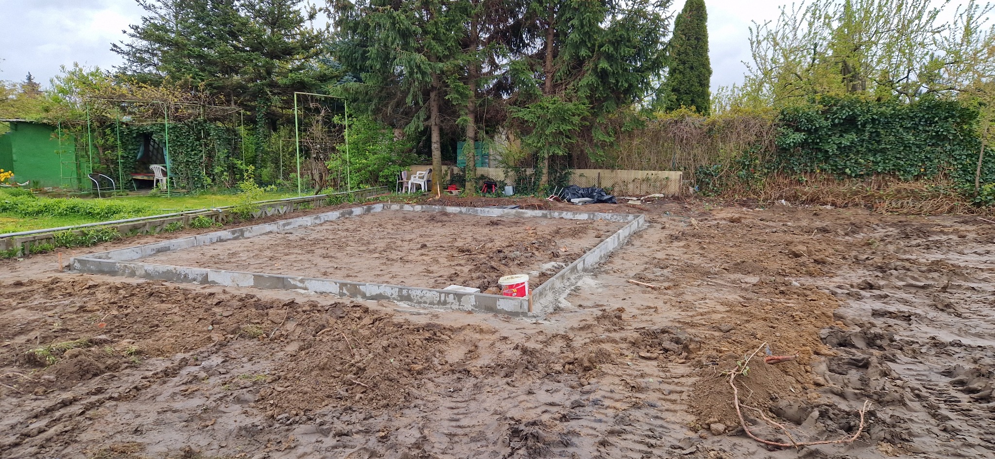 Fundament pod budynek w ogrodzie, z widocznymi betonowymi ścianami i przygotowanym terenem. W tle zieleń drzew i krzewów, pochmurne niebo.