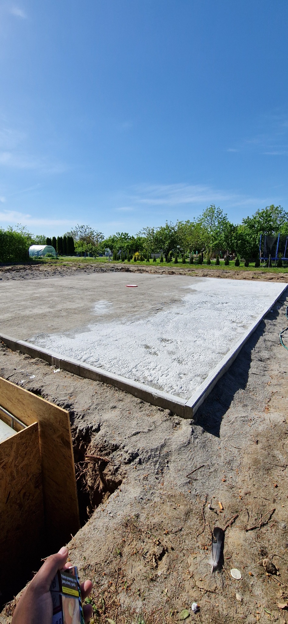 Przygotowany teren pod budowę z wylaną podbudową, obrzeżami i wykopem. Widok na błękitne niebo i zieleń w tle. Ujęcie z dołu, z widoczną ręką trzymającą paczkę papierosów.