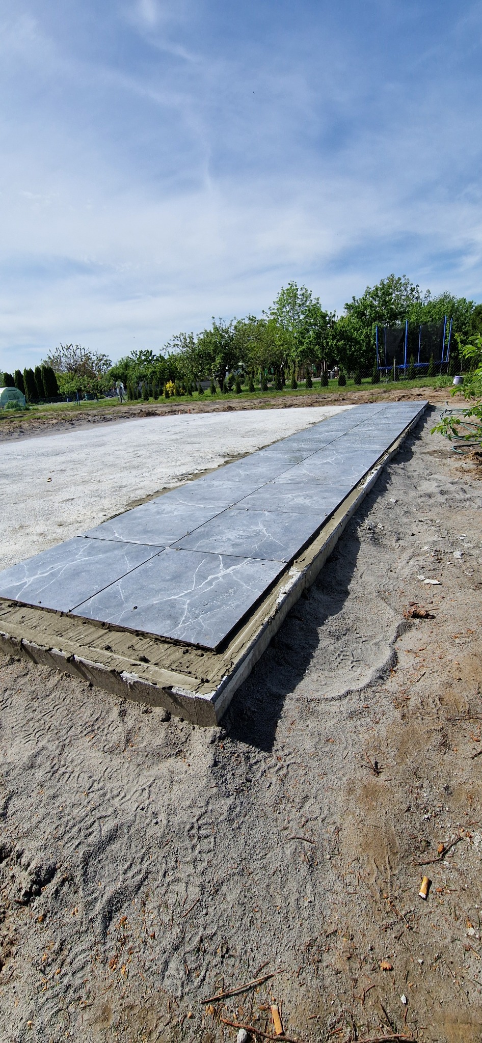 Układanie szarych płyt chodnikowych z marmurkowym wzorem na betonowej wylewce, widok z góry, w tle zieleń ogrodu i błękitne niebo.