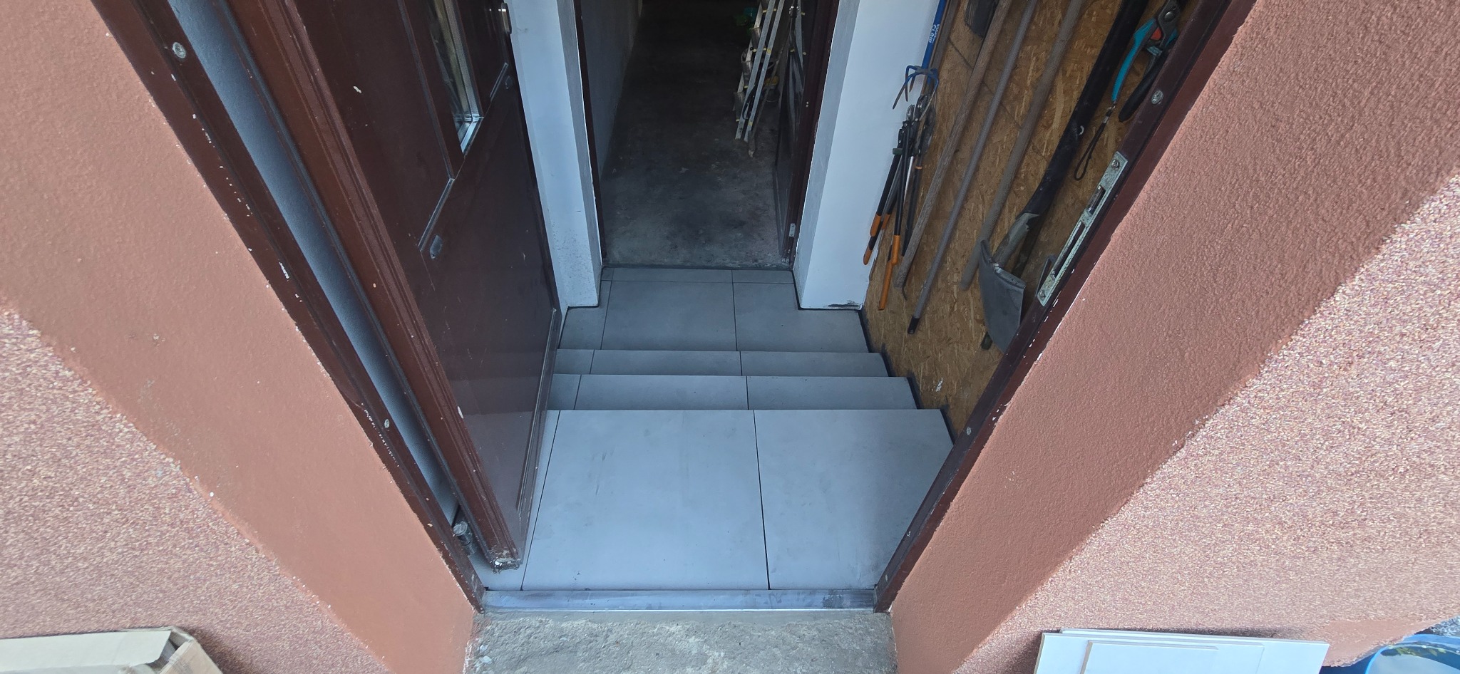 Wykończone schody z szarymi płytkami prowadzące w dół, widoczne narzędzia na ścianie i fragment otwartych, brązowych drzwi wejściowych.