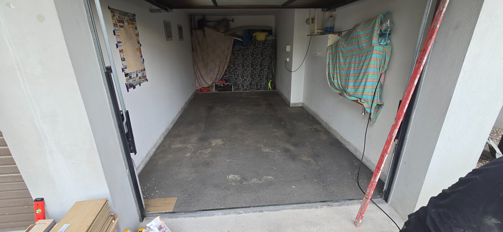 Wnętrze garażu po wylewce betonowej, widoczne narzędzia i poziomica. Ściany białe, na jednej z nich plakat. Podłoże szare, surowe. Otwarta brama garażowa.