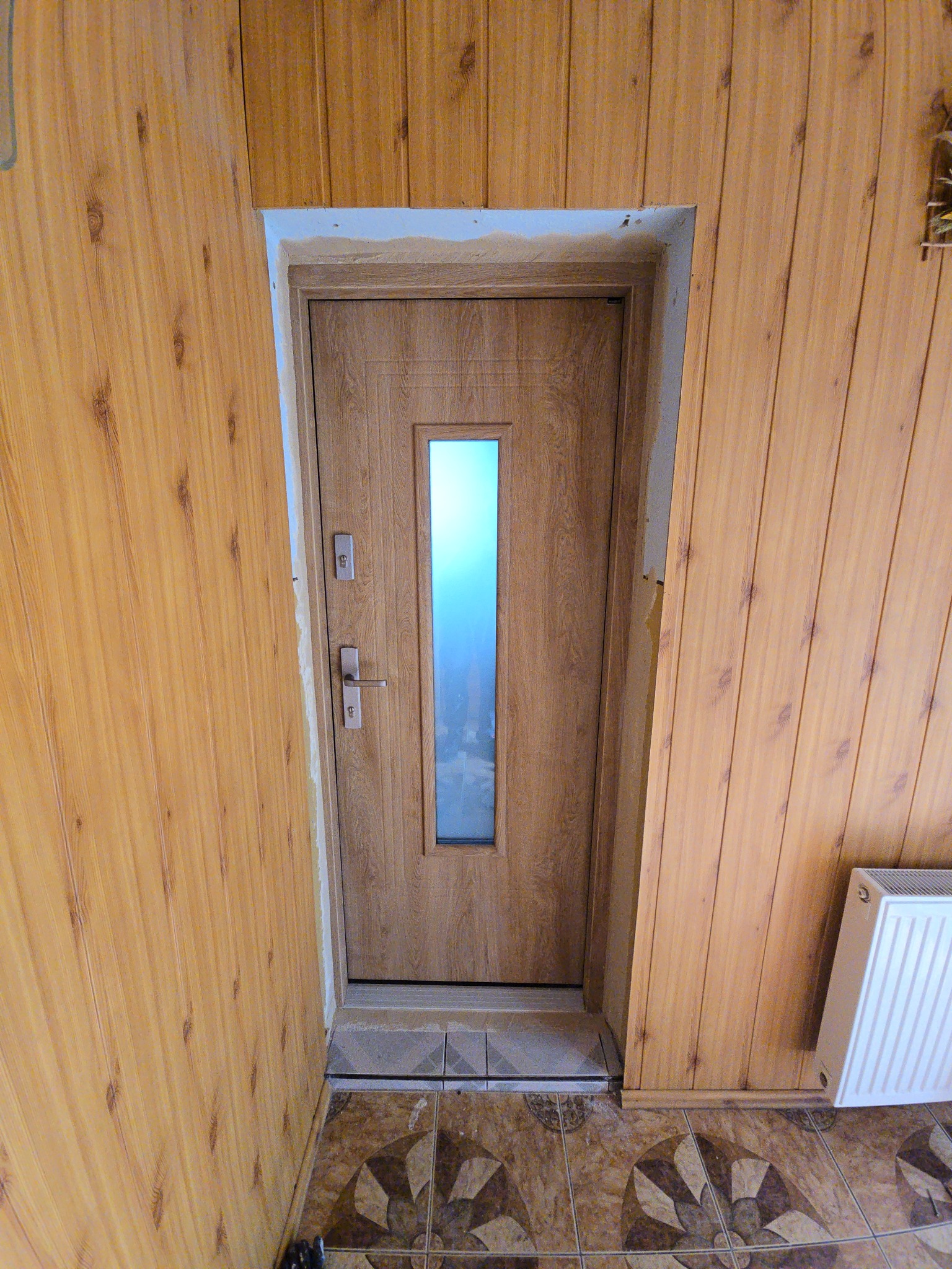 Montaż drzwi wewnętrznych z szybą w obudowie z paneli ściennych. Widoczne wykończenia i fragment grzejnika. Ułożone płytki podłogowe.