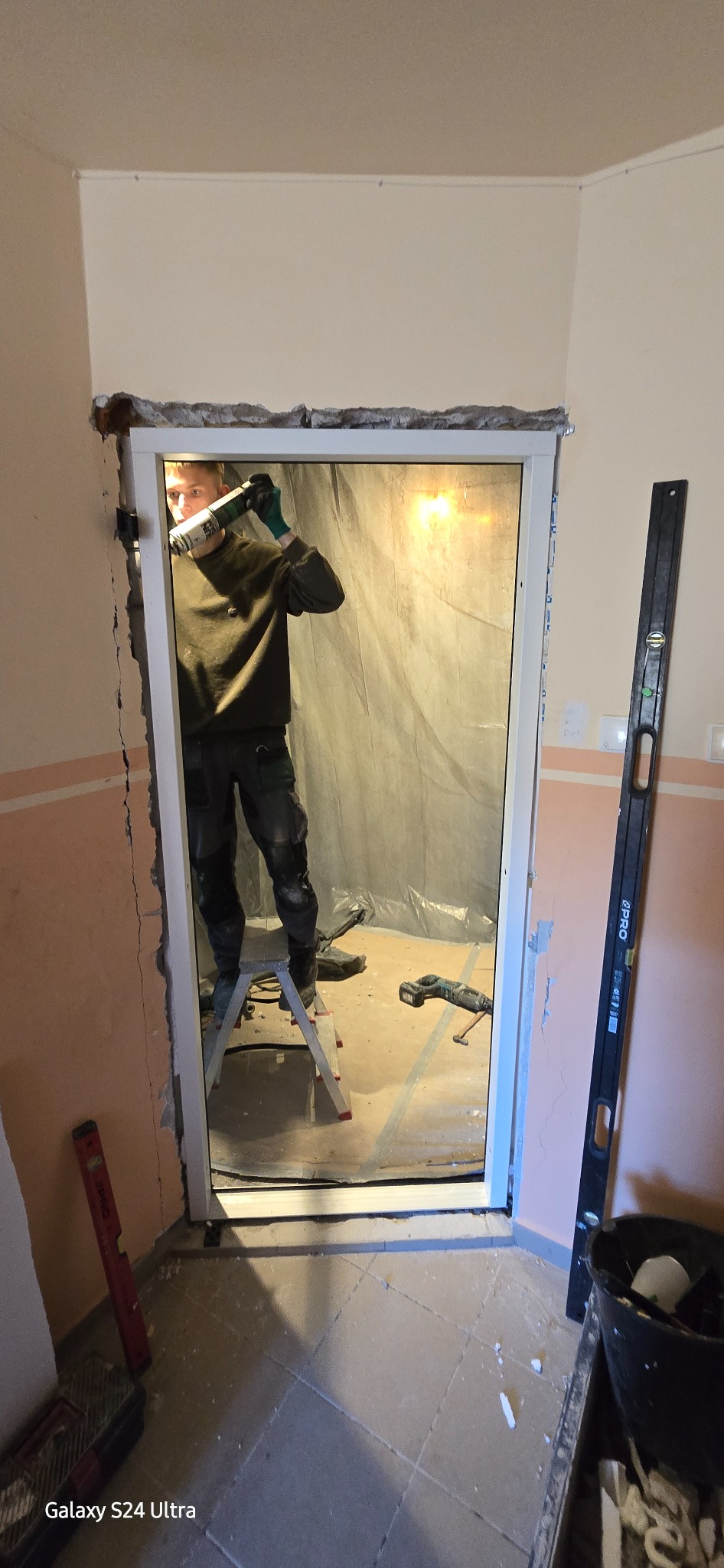 Montaż ościeżnicy: mężczyzna na drabinie uszczelnia pianką ramę drzwiową. Poziomica oparta o ścianę, widoczne narzędzia i zabezpieczenie folią w tle.