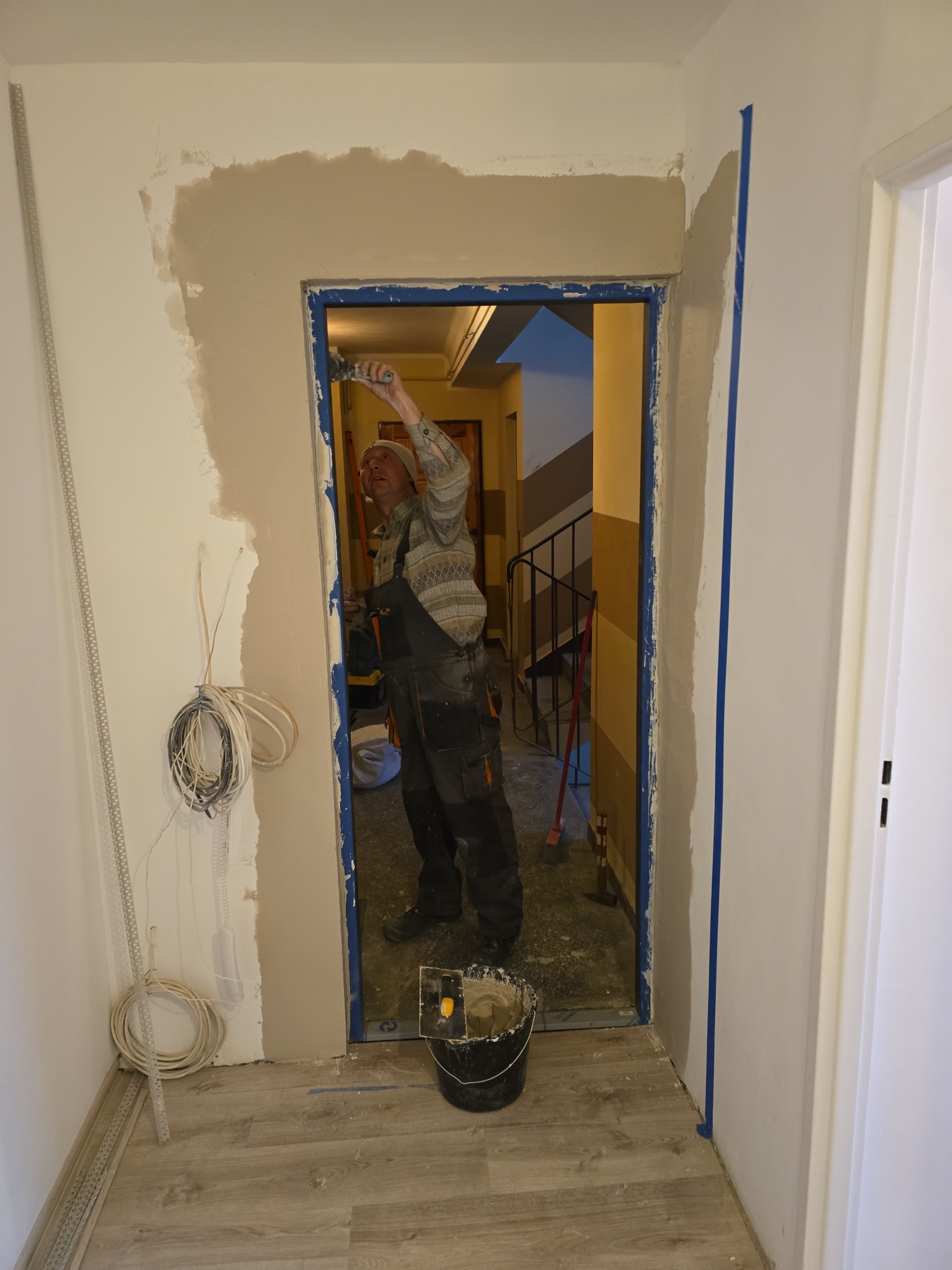 Pracownik w kombinezonie nakłada gładź na ścianę przy futrynie drzwiowej. Widoczne narzędzia, wiązka kabli i fragment klatki schodowej w tle. Prace wykończeniowe.