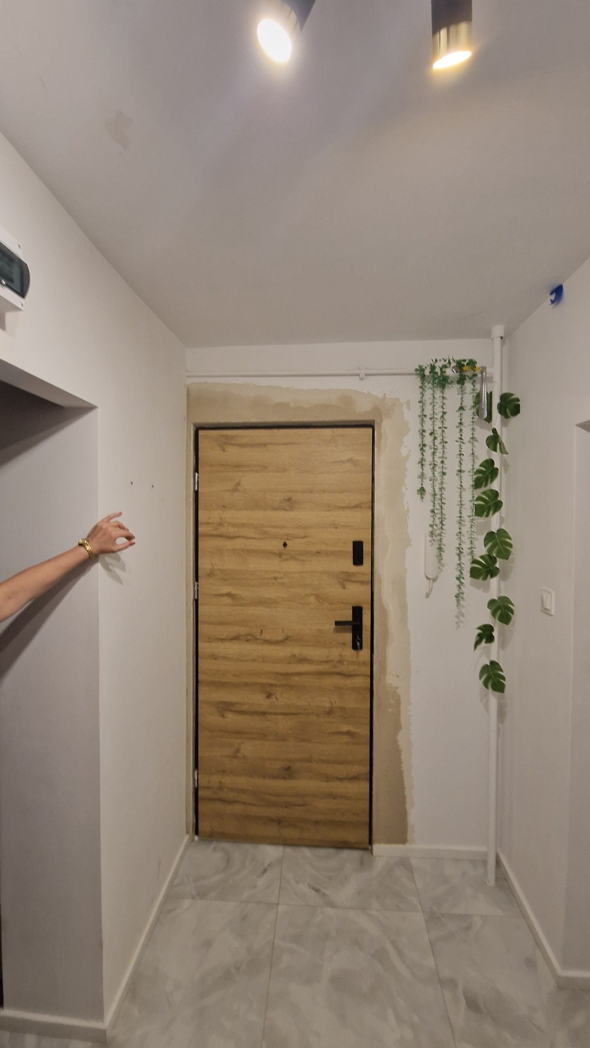 Montaż drzwi wejściowych z drewnianym wzorem, czarne okucia, widoczne ślady po montażu na ścianie, szare płytki podłogowe i dekoracja roślinna.