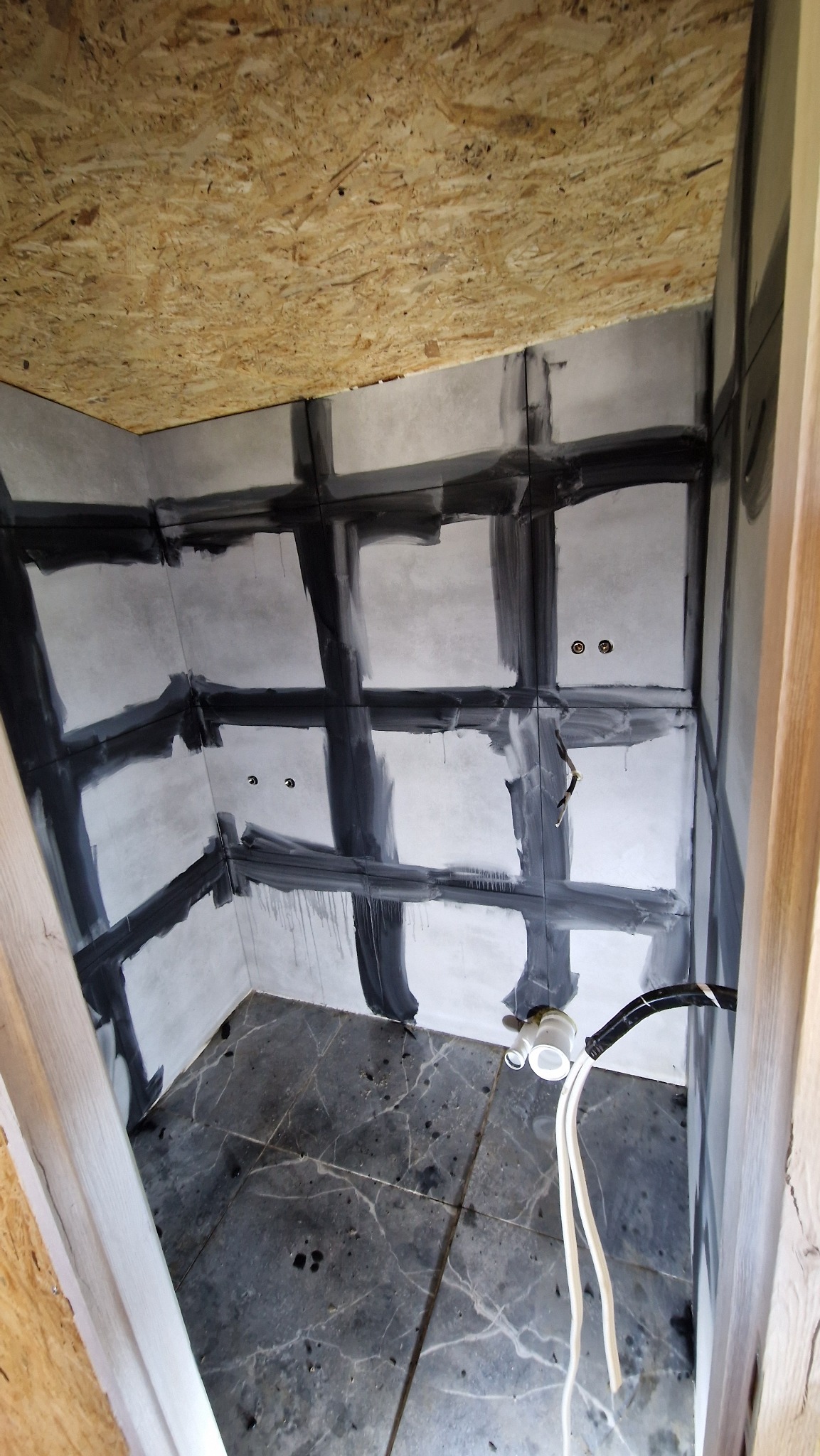 Łazienka w trakcie remontu: szare płytki na ścianach z ciemnymi fugami, marmurowe płytki na podłodze, widoczne instalacje. Sufit wykończony płytą OSB.