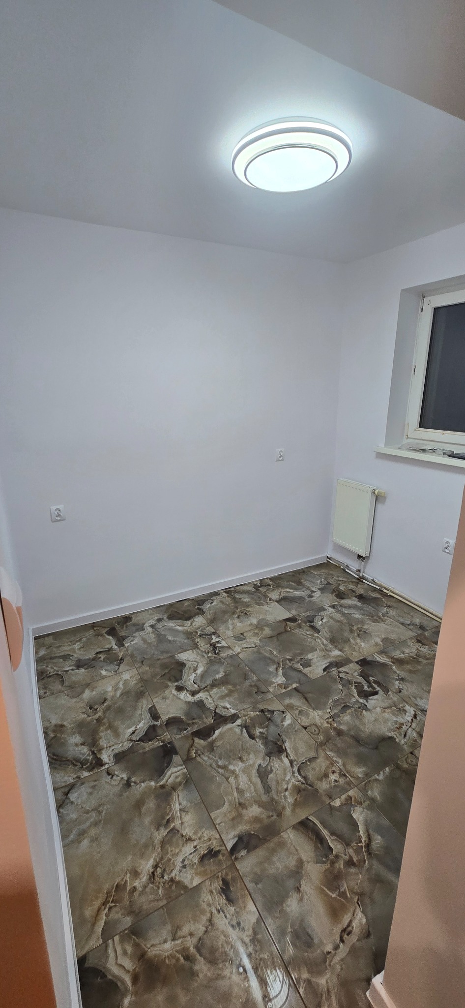 Pokój po remoncie z nową, błyszczącą podłogą z płytek imitujących marmur w odcieniach brązu i beżu. Białe ściany, grzejnik i okno z białymi ramami.