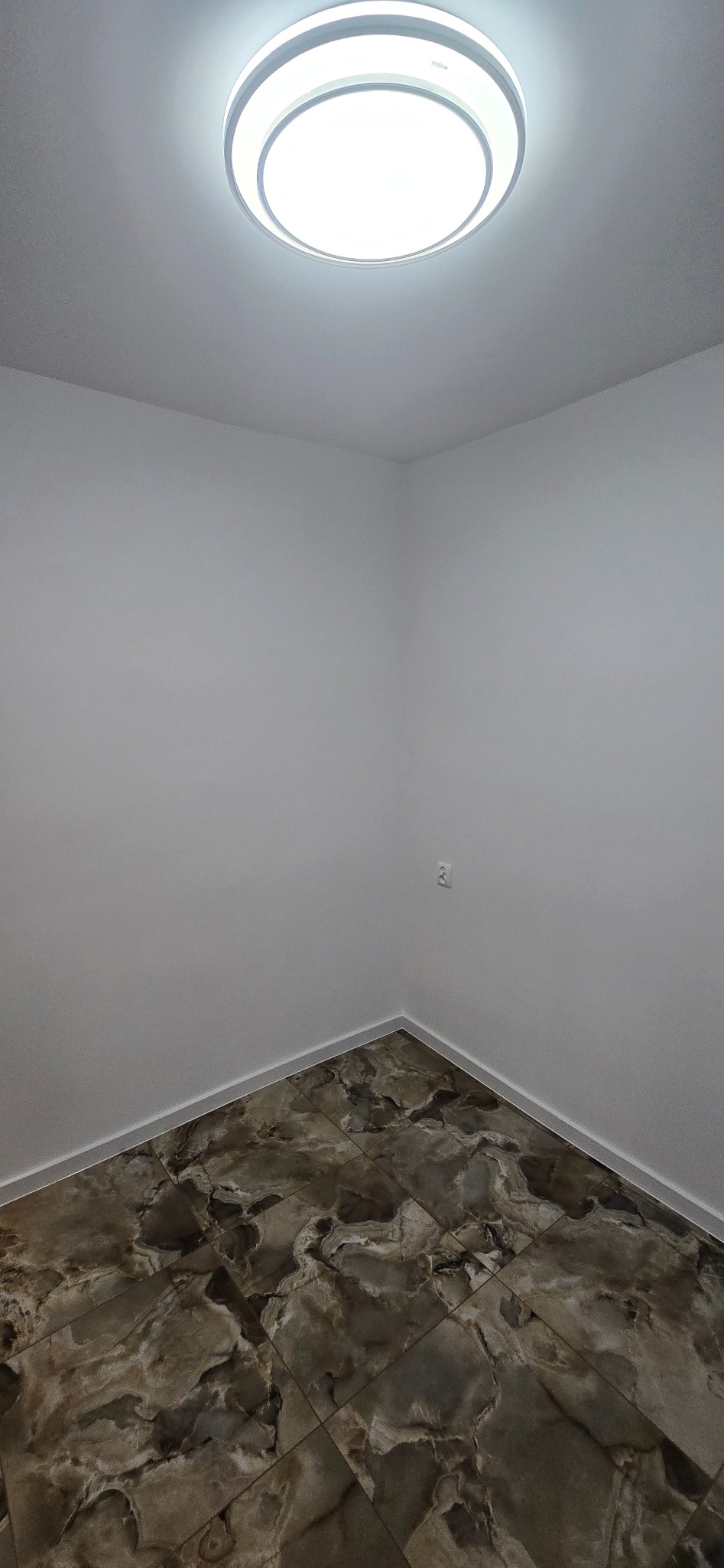 Pusty pokój po remoncie: marmurowe płytki podłogowe w odcieniach brązu i beżu, białe ściany z gładzią i nowoczesna okrągła lampa sufitowa. Widok z góry.