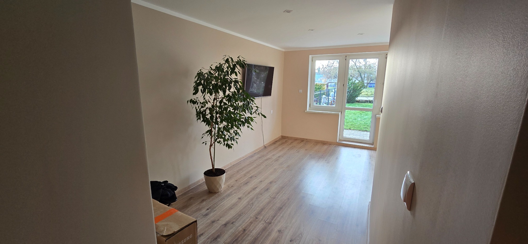 Jasne wnętrze po remoncie: panele podłogowe, ściany w odcieniach beżu, okno z widokiem na ogród, telewizor na ścianie i roślina doniczkowa. Minimalistyczny wystrój.