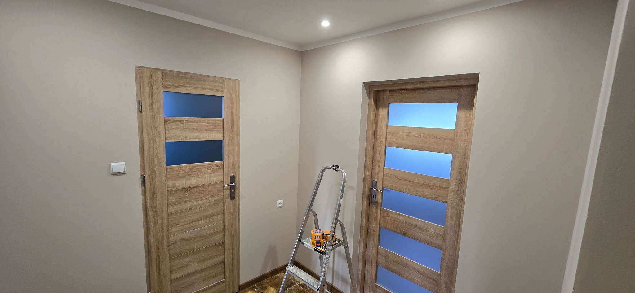 Dwa zamontowane drzwi z przeszkleniami w odcieniach drewna, ustawione w jasnym korytarzu. Obok drabina malarska z puszkami farb. Widoczne listwy przypodłogowe.