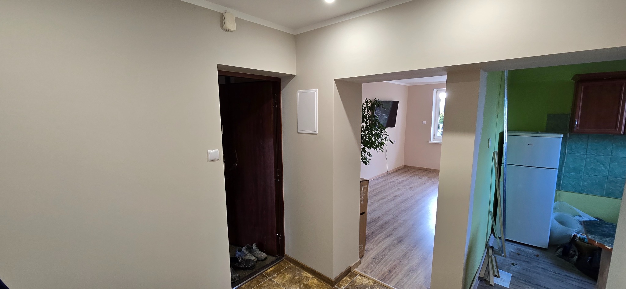 Wykończone wnętrze mieszkania z widokiem na przedpokój z ciemnymi drzwiami, salon z telewizorem i kuchnię z lodówką. Neutralne kolory ścian, panele podłogowe.