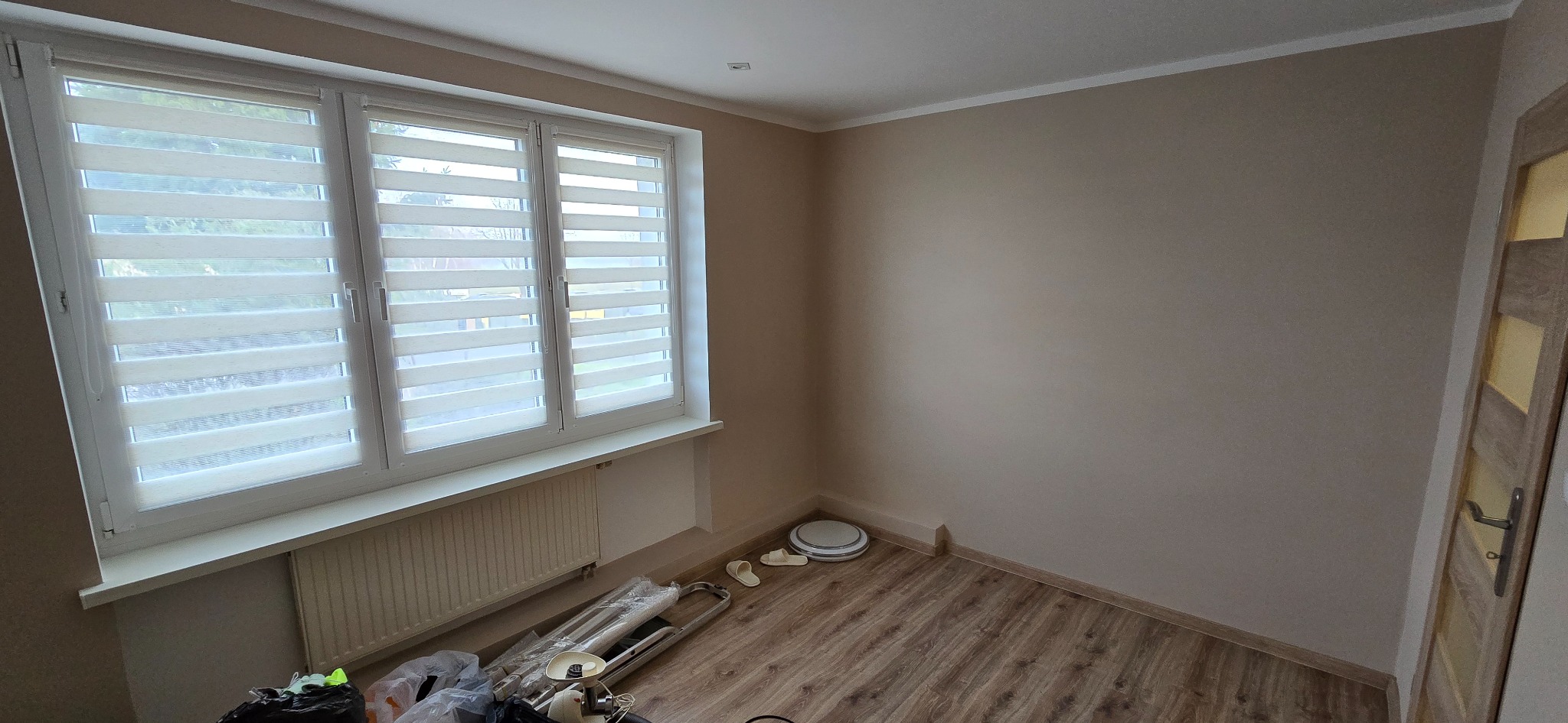Jasne wnętrze pokoju po remoncie: świeżo pomalowane ściany w odcieniach beżu, nowe panele podłogowe i okno z roletami dzień-noc. Widoczne narzędzia i akcesoria malarskie.