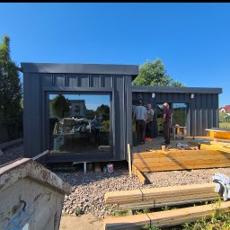 Domy Szkieletowe Mateusz Roszkowski - Montaż nowoczesnego, grafitowego domu modułowego z dużymi oknami. Widoczni pracownicy i materiały budowlane. Słoneczny dzień, błękitne niebo, dom w trakcie budowy.