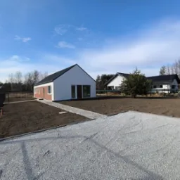 Nowoczesny, parterowy dom z białymi ścianami i antracytowym dachem, otoczony świeżo przygotowanym terenem pod trawnik i żwirowym podjazdem. Błękitne niebo z delikatnymi chmurami.