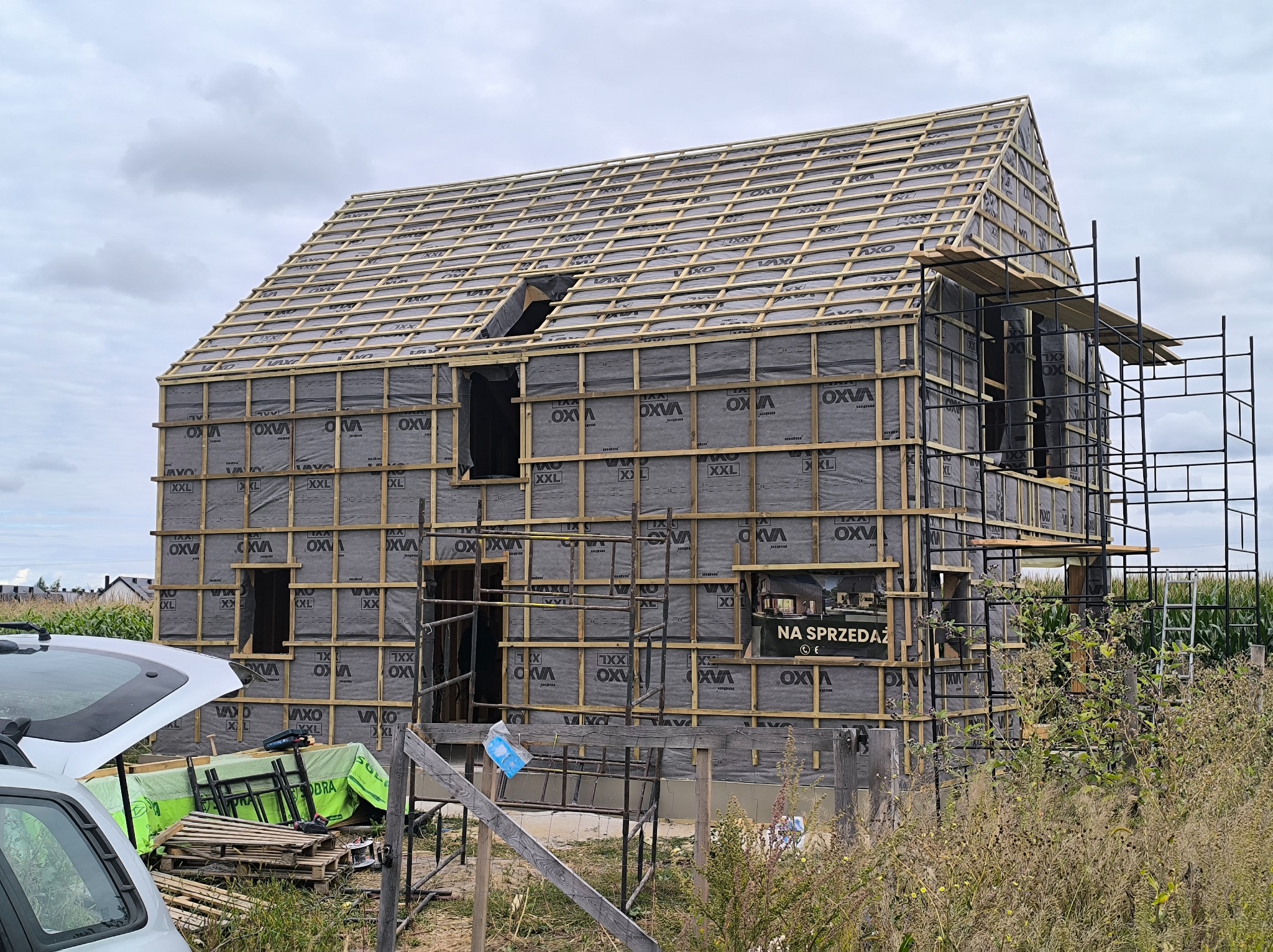 Budowa domu w stanie surowym otwartym: widoczne drewniane rusztowanie dachu, ściany pokryte folią i częściowo obudowane drewnem. Tablica 'Na sprzedaż' na elewacji.