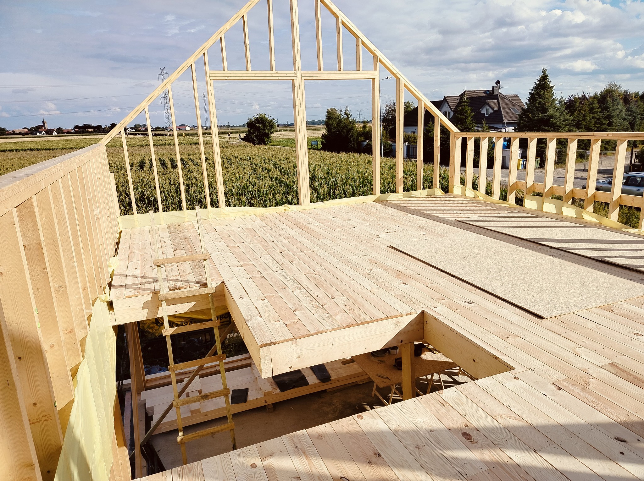 Wnętrze domu szkieletowego w budowie, widok z góry na drewnianą konstrukcję, z zielonym polem w tle i elementami dachu oraz schodami.