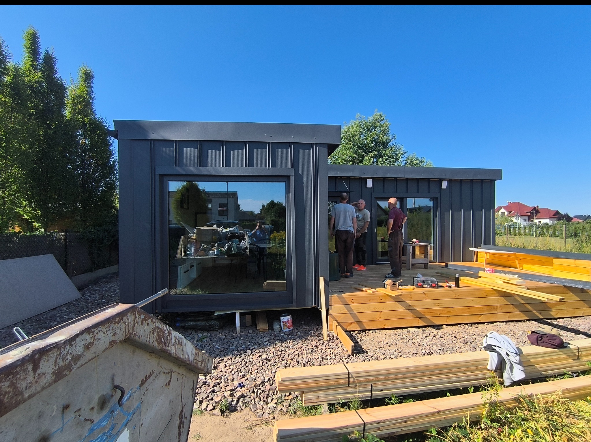 Montaż nowoczesnego, grafitowego domu modułowego z dużymi oknami. Widoczni pracownicy i materiały budowlane. Słoneczny dzień, błękitne niebo, dom w trakcie budowy.