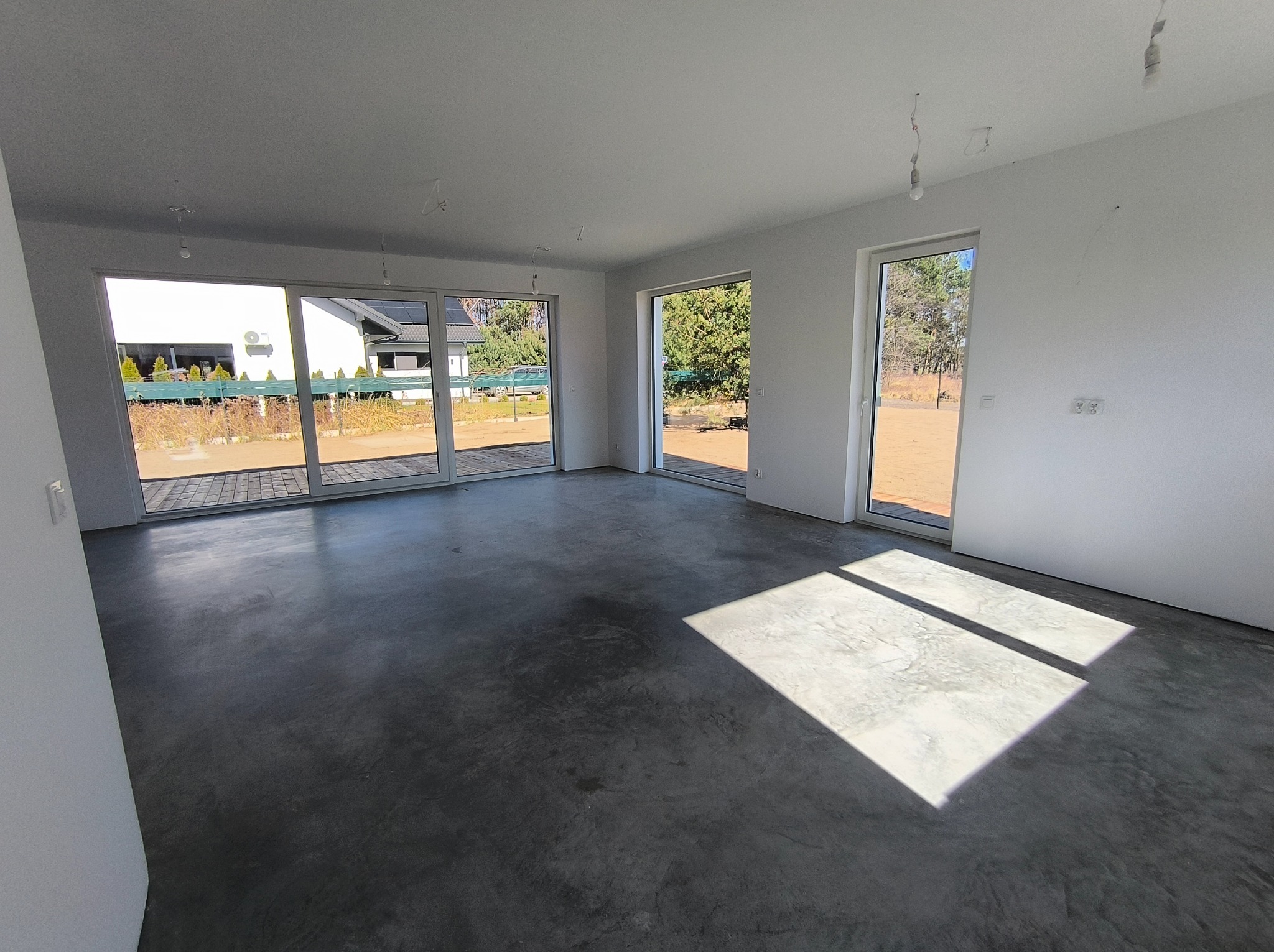 Wnętrze domu w stanie deweloperskim z widoczną wylewką podłogową i dużymi oknami z widokiem na zewnątrz. Na suficie widoczne wystające kable.