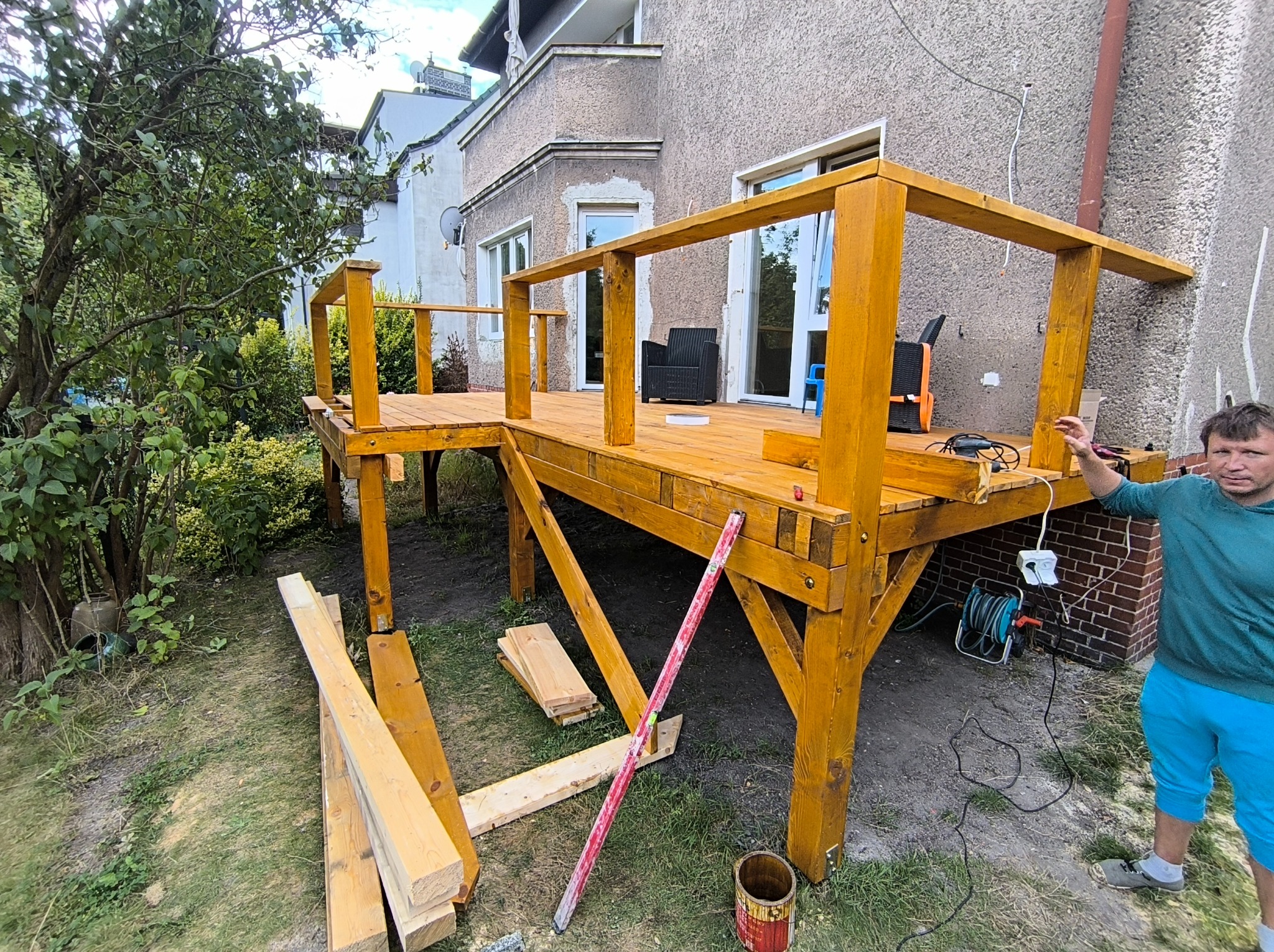 Drewniany taras przy domu w trakcie budowy, widoczne elementy konstrukcyjne, poziomica i osoba. Taras w kolorze miodowym, z balustradą. Częściowo zadaszony.