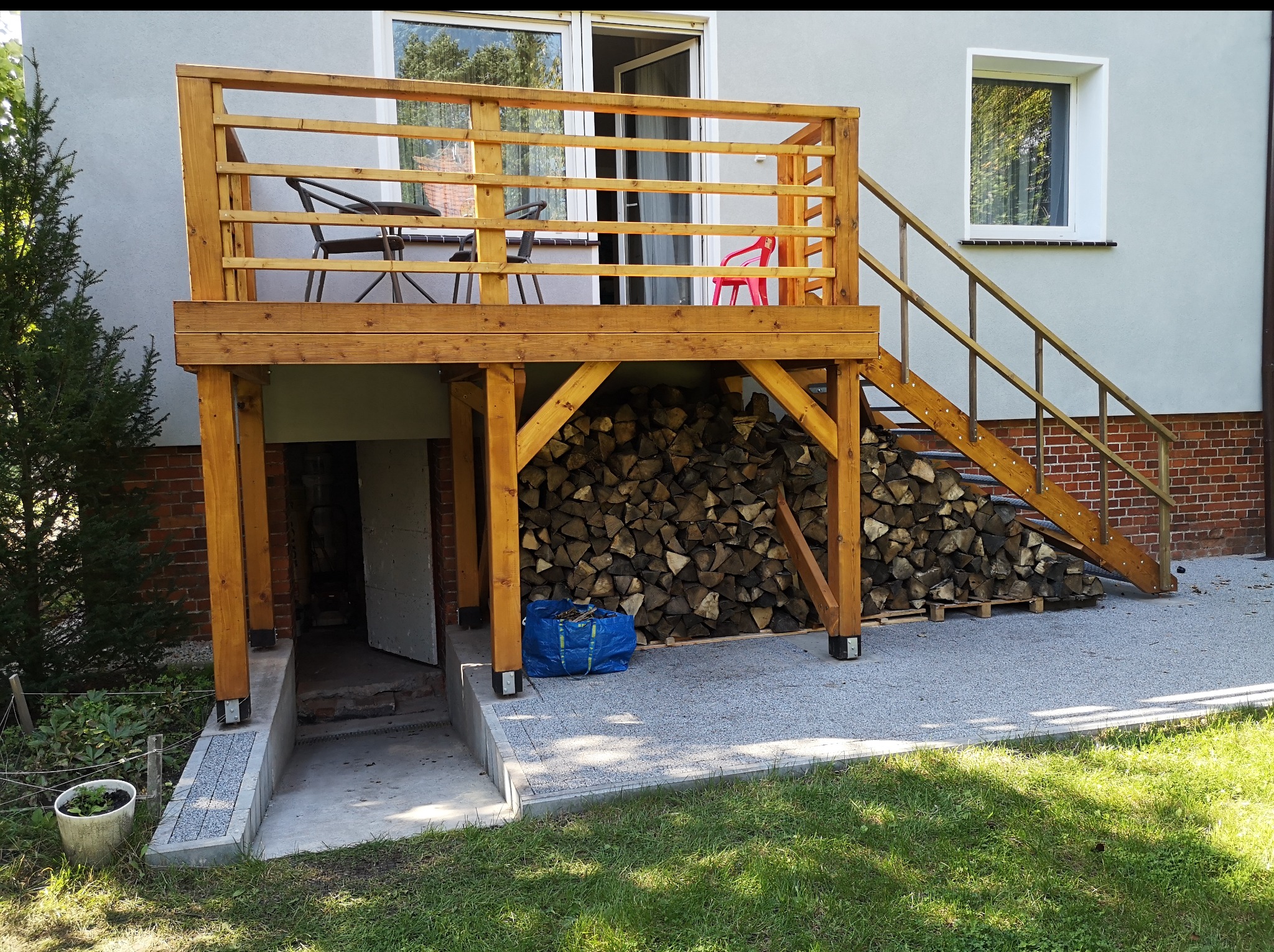 Drewniany taras z barierkami i schodami przy domu. Pod tarasem składowane drewno. Wejście do piwnicy. Widok na okno i drzwi balkonowe.
